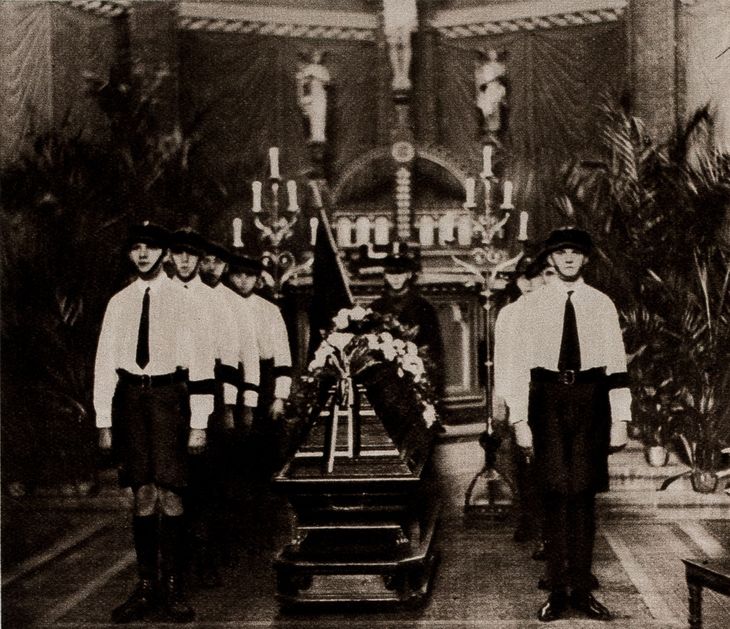 Historisches Schwarz-Weiß-Foto: neben einem mit Blumen geschmückten Sarg stehen acht junge Männer mit Schirmmützen in einer Kapelle.