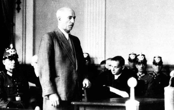 Historisches Schwarz-Weiß-Foto: Weilhelm Leuschner steht an einem kleinen Tisch in einem Gerichtssaal und hält sich an dem vor ihm stehenden Stuhl fest. Hinter ihm sitzen Polizisten.