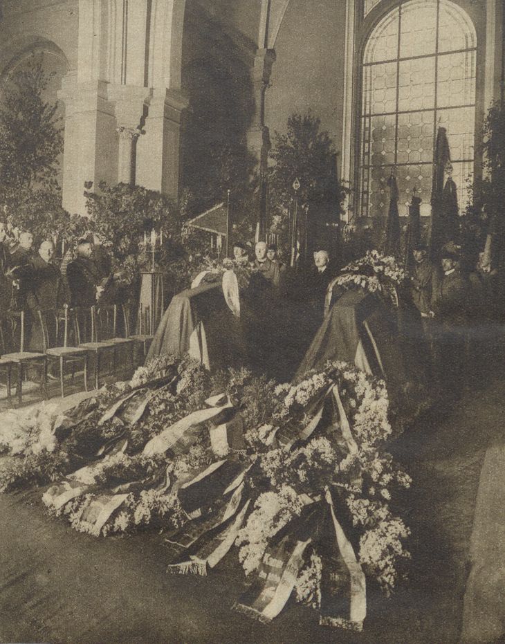 Historisches Schwarz-Weiß-Foto von einer Kapelle mit zwei Särgen und Kränzen. Im Hintergrund stehen mehrere Männer.