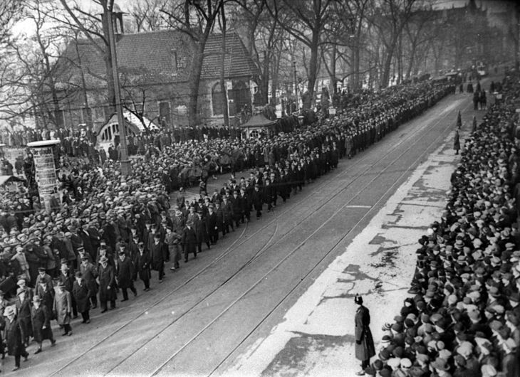 Historisches Schwarz-Weiß-Foto: über eine breite Straße zieht ein langer Menschenzug. Auf dem Gehsteig stehen dicht gedrängt zahlreiche Passanten.