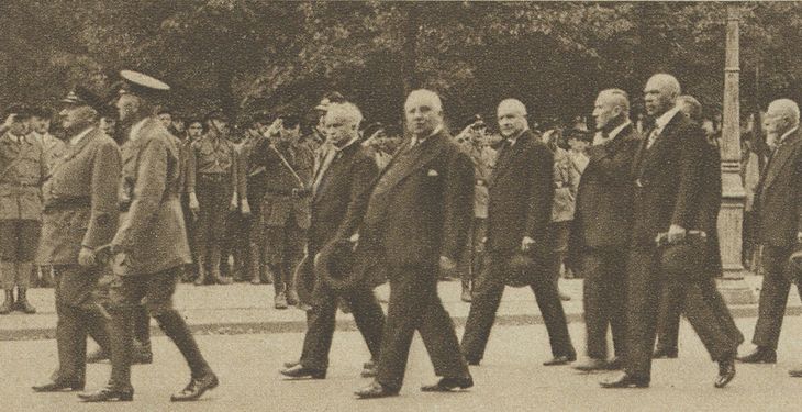 Historisches Schwarz-Weiß-Foto: Männer in Anzug und mit Hut in der Hand gehen eine Straße entlang. Auf dem Gehsteig stehen Reichsbannermänner und salutieren.