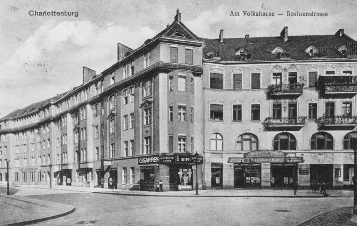 Historische Postkarte mit einem Schwarz-Weiß-Foto eines vierstöckigen Wohnhauses mit einer Ladenzeile. Darüber steht: Charlottenburg, Am Volkshause - Rosinenstrasse.