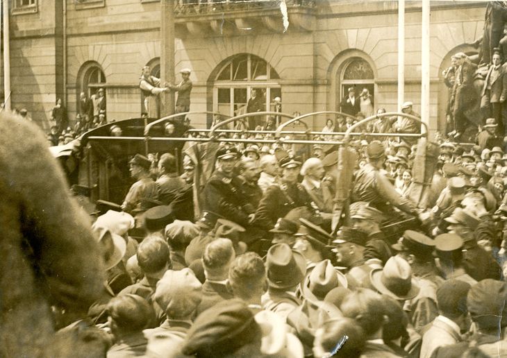 Historisches Schwarz-Weiß-Foto: Männer werden von der Polizei begleitet auf einem offenen Wagen durch eine Menschenmenge gefahren.