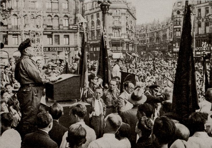 Historisches Schwarz-Weiß-Foto: Ernst Mulansky steht an einem Rednerpult uns spricht zu einer großen Menschenmenge auf einem Platz vor mehrstöckigen Wohnhäusern.