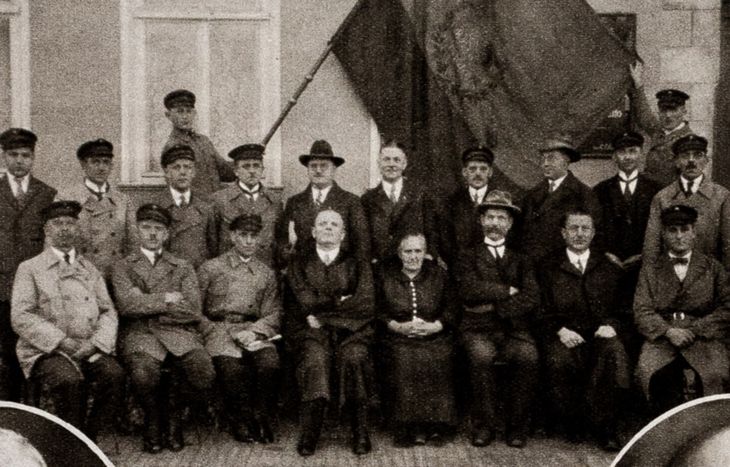 Historisches Schwarz-Weiß-Foto: Gruppenaufnahme von 19 Männern und einer Frau vor einem Gebäude. Die Männer tragen Anzüge oder Reichsbanner-Uniformen, die Frau ein Kleid. Zwei der Männer stehen im Hintergrund und halten eine große Fahne mit Reichsadler. 