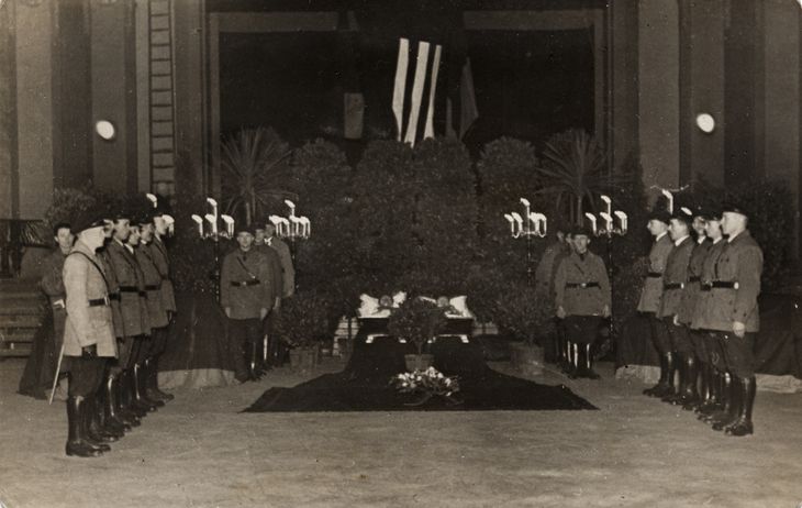 Historisches Schwarz-Weiß-Foto: neben einem mit Kränzen geschmückten Sarg stehen in vier Reicgeb junge Männer in Reichsbanner-Uniformen und mit Schirmmützen. Im Hintergrund ist eine Fahne mit den drei Pfeilen der Eisernen Front zu erkennen.