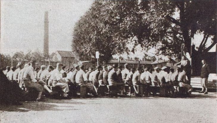 Historisches Schwarz-Weiß-Foto: Auf einem Platz sitzen Männer auf hintereinander aufgereihten Bänken vor einem Rednerpult. An dem Rednerpult steht ein Mann und spricht, daneben ein weiterer Mann, der die Arme hinter dem Rücken gekreuzt hat. Hinter dem Rednerpult sind zwei Fahnen angebracht..