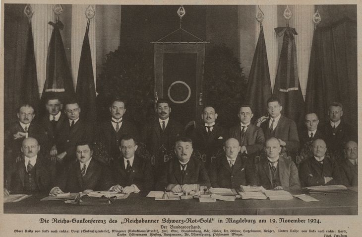 Historischer Zeitungsausschnitt mit einem Schwarz-Weiß-Foto: 18 Männer sitzen bzw. stehen in zwei Reihen an einem langen Tisch voller Dokumente. Hinter ihnen sind sieben Fahnen zu sehen.
